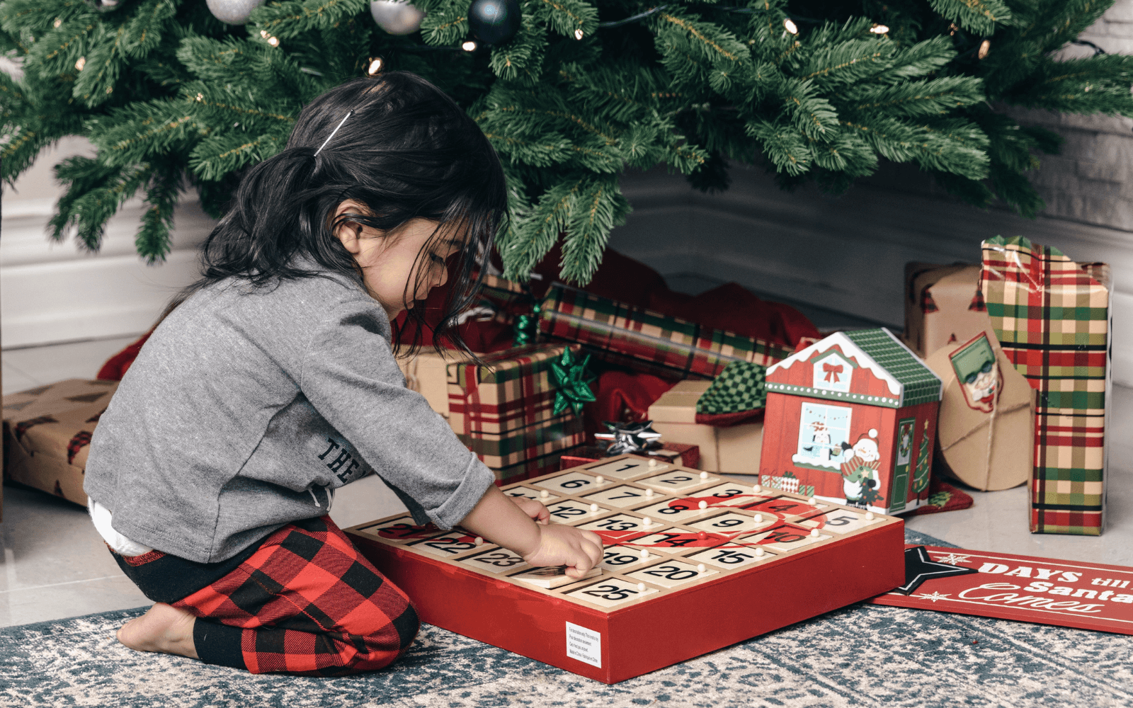 Child opening a numbered wooden Advent calendar beneath a decorated Christmas tree, surrounded by wrapped gifts.