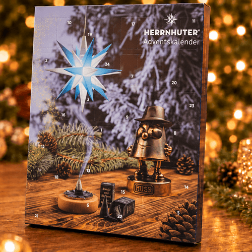 A HERRNHUTER Adventskalender (Christmas advent calendar) standing on a wooden table with a festive blurred background of Christmas lights and pine branches. The calendar cover features a star-shaped ornament, a small metal gnome, and numbered compartments.