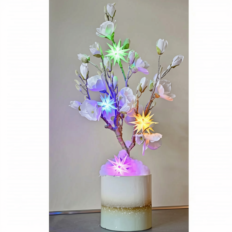 An illuminated artificial flowering tree with white blossoms and glowing multicolor starburst lights, displayed in a cylindrical white pot with a gold glitter band, set against a plain beige wall.