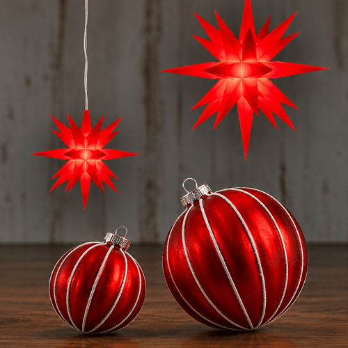 Two Herrnhut Stars in red on a dark wooden surface against a rustic gray with red silver ornaments.